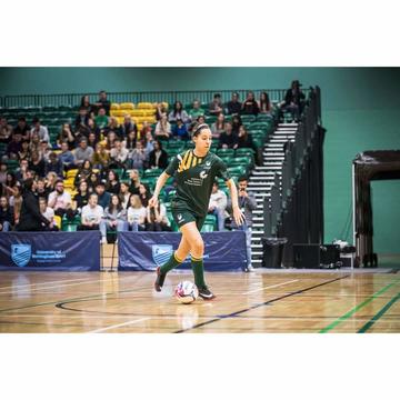 Morgan plays football in an indoor gymnasium. Wide shot of her with a ball and a crowd in the background. She is wearing green kit. 